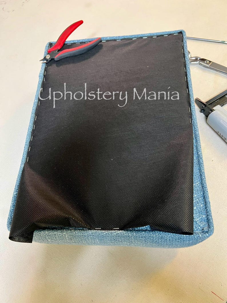 May include: A close-up of a blue fabric-covered cushion with a black fabric lining. The cushion is being stapled together with a staple gun. The text "Upholstery Mania" is visible on the cushion.