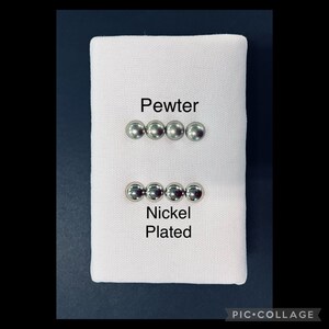 May include: A white fabric square displays two sets of four small, round, metallic objects. The top row is labelled "Pewter," and the bottom row is labelled "Nickel Plated." The objects are likely buttons or decorative elements.