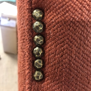 May include: A close-up shot of a rust-coloured blazer, showcasing a textured herringbone weave. The blazer's edge features six ornate, gold-toned buttons. The buttons' intricate design complements the fabric's pattern, adding a classic touch to the garment.