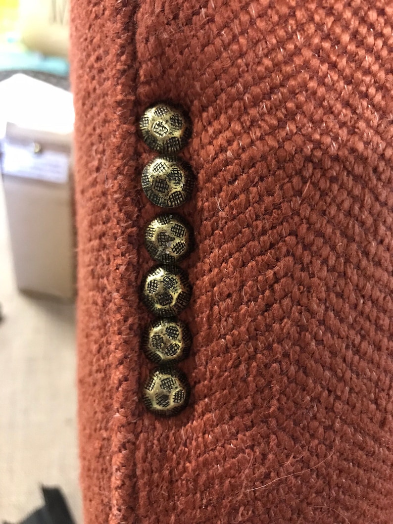 May include: A close-up shot of a rust-coloured blazer, showcasing a textured herringbone weave. The blazer's edge features six ornate, gold-toned buttons. The buttons' intricate design complements the fabric's pattern, adding a classic touch to the garment.