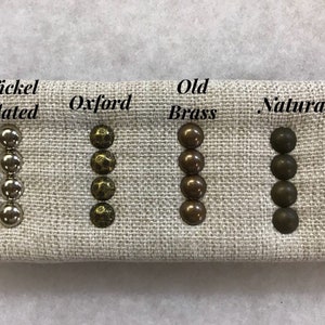 May include: A fabric sample shows four sets of upholstery tacks in different finishes: Nickel Plated, Oxford, Old Brass, and Natural. The tacks are arranged in vertical rows of four, displaying their varied colours and textures. Text labels identify each finish.