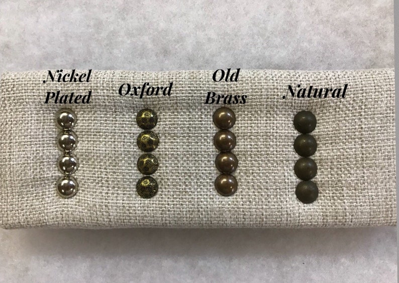 May include: A fabric sample shows four sets of upholstery tacks in different finishes: Nickel Plated, Oxford, Old Brass, and Natural. The tacks are arranged in vertical rows of four, displaying their varied colours and textures. Text labels identify each finish.