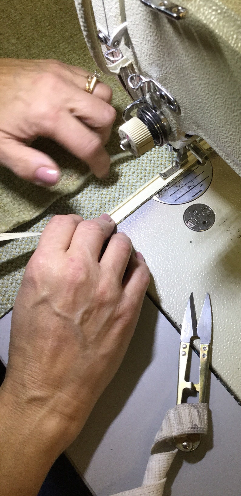 May include: A close-up of a sewing machine with a beige fabric being fed through the needle. A hand is holding the fabric in place. A pair of small silver scissors with a beige fabric wrapped around the handles is on the table.