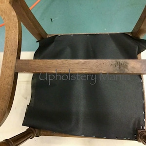 May include: A close-up of a wooden chair with a black fabric seat. The chair is being reupholstered. The text "Upholstery Mania" is visible on the seat.