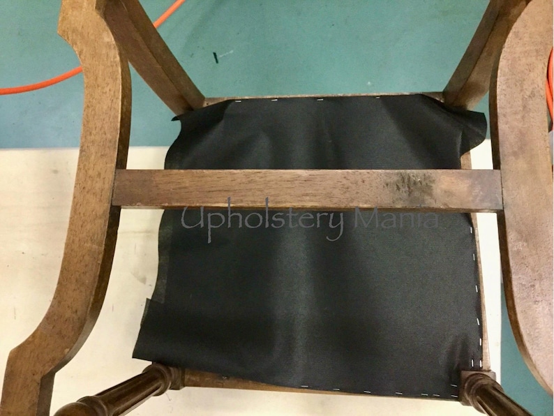 May include: A close-up of a wooden chair with a black fabric seat. The chair is being reupholstered. The text "Upholstery Mania" is visible on the seat.