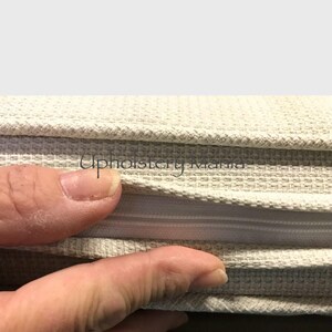 May include: Close-up of a beige fabric with a zipper. The fabric is folded over to show the zipper. The zipper is white and has a metal slider.
