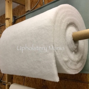 May include: A white roll of upholstery batting, a type of fabric used for padding and cushioning, is rolled up and secured with a wooden dowel. The roll is labeled "Upholstery Mania".