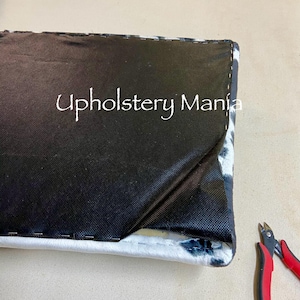 May include: Close-up of a black and white patterned fabric being sewn with a black thread. A pair of red wire cutters are on the right side of the image. The text "Upholstery Mania" is visible on the fabric.