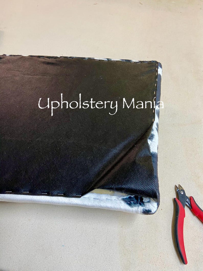 May include: Close-up of a black and white patterned fabric being sewn with a black thread. A pair of red wire cutters are on the right side of the image. The text "Upholstery Mania" is visible on the fabric.