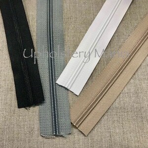 May include: Four different zipper tapes, black, gray, white, and beige, laid out on a beige fabric background.