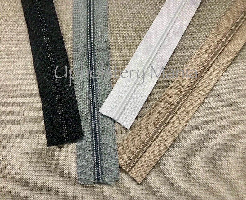 May include: Four different zipper tapes, black, gray, white, and beige, laid out on a beige fabric background.