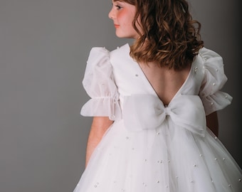 May include: A white tulle dress with a large bow on the back and scattered pearl accents. The dress features short, puffy sleeves and a V-neckline. A pearl headband adorns the hair. The dress is suitable for special occasions.