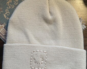 Puede incluir: Gorro de punto beige con borde doblado. Un parche cuadrado cosido con una cara sonriente está en el borde. El gorro está hecho de un material suave y texturizado, perfecto para el invierno.