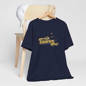 May include: Navy blue t-shirt featuring the word "Taurus" in a retro yellow and blue font, with a constellation design. The shirt is hanging on a wooden hanger, with a white cap resting on a wooden chair in the background.