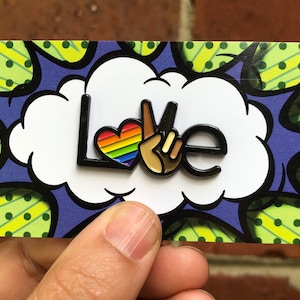 May include: A black and white enamel pin with the word "Love" in a comic book style font. The "O" in Love is replaced with a rainbow heart and a peace sign.