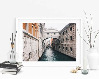 Puede incluir: Una fotografía enmarcada de un canal estrecho en Venecia, Italia. El canal está bordeado de edificios a ambos lados, y un puente se arquea sobre el agua. El agua está tranquila y reflectante, y los edificios son una mezcla de blanco y naranja.