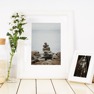 May include: Two white picture frames with a white background. The frame on the left shows a stack of rocks on a beach with a body of water in the background. The frame on the right shows a forest scene. The frames are on a wooden surface with a vase of white flowers and a glass jar.