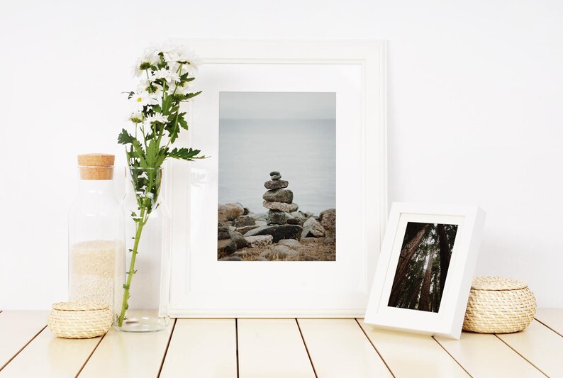 May include: Two white picture frames with a white background. The frame on the left shows a stack of rocks on a beach with a body of water in the background. The frame on the right shows a forest scene. The frames are on a wooden surface with a vase of white flowers and a glass jar.