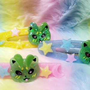 May include: Four colorful hair clips with glittery accents. Each clip features a different color: yellow, blue, and pink. The clips are decorated with star and moon shapes, and a green cat face. The background is a rainbow of colors.