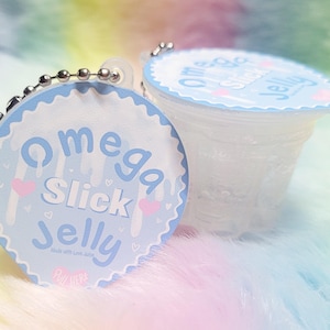May include: A small, clear, cup-shaped container with a light blue circular lid featuring the text Omega Slick Jelly, small pink hearts, and white drip patterns. A silver metal ball chain is attached to the top edge of the lid for hanging.