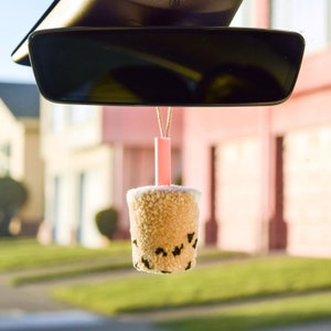 May include: A brown and white boba tea-shaped car air freshener with a pink straw hanging from the rearview mirror of a car.