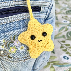 May include: A yellow crocheted star-shaped charm with black bead eyes and a smiling mouth. The charm is attached to a yellow braided cord. The background includes a denim pocket with floral embroidery.