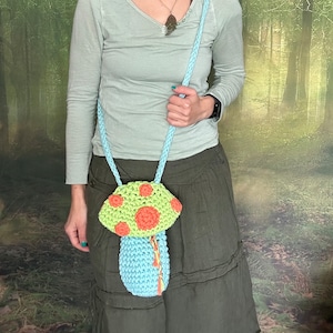 May include: A crocheted mushroom-shaped bag with a light blue body, green and orange accents, and a long strap. The bag is worn by a person in a forest setting.