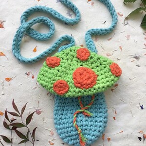 Puede incluir: Una bolsa de ganchillo con forma de hongo con una correa azul claro. El hongo es verde con lunares naranja y un tallo azul claro.