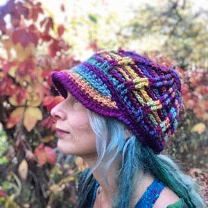 May include: A colorful crocheted hat with a wide brim. The hat is made with a variety of colors, including purple, blue, green, yellow, and orange. The hat is worn by a person with blue hair.