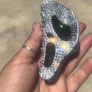 May include: A silver-colored key fob covered in small, sparkling rhinestones. The key fob has two black, oval-shaped accents and a green accent. The rhinestones reflect light, creating a dazzling effect.