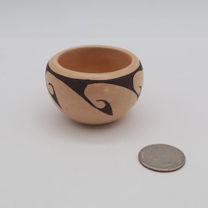 May include: A small, round, beige ceramic bowl with a dark brown, swirling design around the rim. The bowl is empty and sits on a white surface. A coin is next to the bowl.