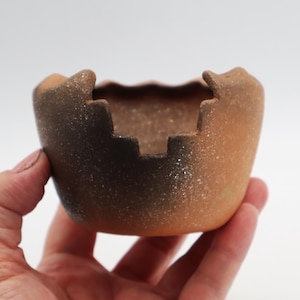 May include: A small, handcrafted ceramic bowl with a unique, irregular rim. The bowl features a gradient of colors, transitioning from a dark brown base to a warm, reddish-brown upper portion. The textured surface adds to its rustic charm.