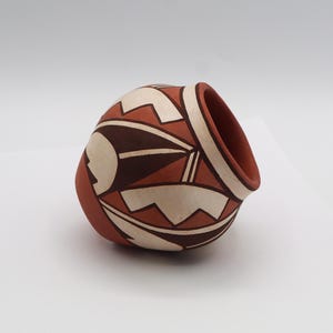 May include: A handmade, terracotta-colored pottery vessel with a wide opening. The exterior features geometric designs in cream and dark brown. The vessel has a rounded shape and is likely used for decorative or functional purposes.