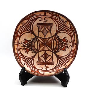 May include: A round, beige ceramic bowl with a detailed geometric design in dark brown and reddish-brown. The bowl is displayed on a black stand, showcasing its intricate patterns and craftsmanship. The design features symmetrical shapes and lines.