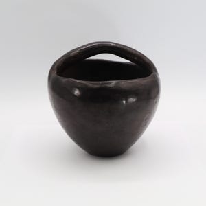 May include: A dark brown ceramic basket with a rounded body and a curved handle. The basket has a smooth, glossy finish and sits on a white surface. The handle is integrated into the design, creating a seamless look.