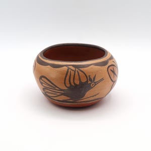 May include: A small, round, earthenware bowl with a reddish-brown interior and exterior. The exterior features a tan background with a black bird design and a scalloped border. The bowl is likely handmade and has a rustic aesthetic.