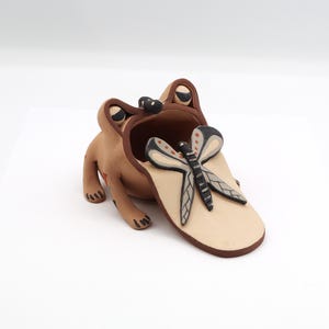 May include: A whimsical ceramic frog-shaped container in shades of brown and tan. A decorative dragonfly with black, gray, and red accents rests on the frog's tongue. The frog has large, expressive eyes and a wide open mouth.