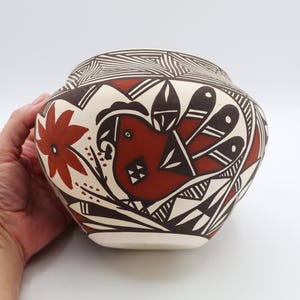 May include: A hand-painted ceramic pot with a rounded shape. The pot features a bird and floral design in shades of red and brown against a cream-colored background. Geometric patterns and lines decorate the upper portion of the pot.