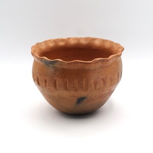 May include: A small, round, terracotta-colored ceramic bowl with a ruffled rim. The bowl has a series of vertical indentations around the middle. The exterior has a slightly rough texture and a few dark spots. The bowl is set against a white background.