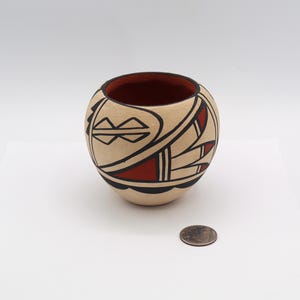May include: A round, beige ceramic pot with a red interior and black and red geometric designs. The pot is sitting on a white surface. A coin is next to the pot.