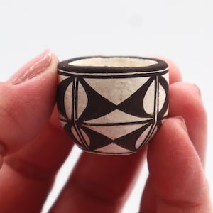 May include: A small, round, hand-painted ceramic bowl. The bowl features a cream-colored base with a repeating geometric pattern in black. The rim is also black. The bowl is held in a person's hand.