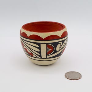 May include: A small, round, hand-painted ceramic bowl with a red interior and cream-colored exterior. The bowl features a black and red geometric design. A quarter is next to the bowl for size comparison.