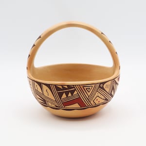 May include: Handmade ceramic basket with a rounded bowl and arched handle. The basket is beige with a geometric pattern in dark brown and red around the rim. The handle also has a decorative pattern.