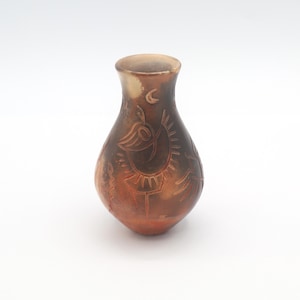 May include: A small, handcrafted ceramic vase with a bulbous body and a narrow neck. The vase is primarily brown with reddish-orange accents and features etched designs of birds and crescent moons. The vase is approximately 5 inches tall.