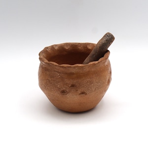 May include: A small, round, unglazed terracotta mortar and pestle set. The mortar has a textured surface with small indentations. The pestle is a dark brown color and is inserted into the mortar. The mortar is approximately 3 inches in diameter.