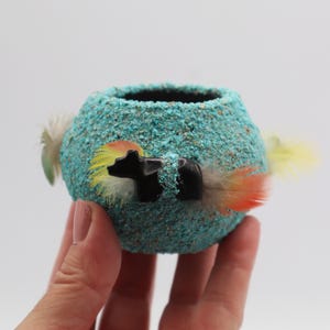 May include: A small, round turquoise-colored pot with a textured surface. The pot features two black bear figures and colorful feathers. The pot is held in a person's hand against a white background.