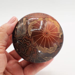 May include: A brown, spherical decorative object with intricate carved floral and geometric patterns. The object has a central circular design and is held in a hand. The color is a mix of dark and light brown.
