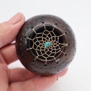 May include: A dark brown, spherical decorative object with a dreamcatcher design. The dreamcatcher is made of a light tan web with a small turquoise stone in the center. The object has a carved pattern around the edge.