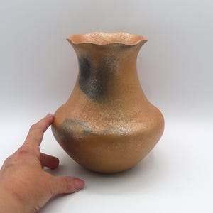 May include: A handcrafted ceramic vase with a ruffled rim. The vase has a warm, earthy color with a speckled texture and a dark gray accent. The vase is a decorative item.