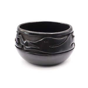 May include: A black, round ceramic bowl with a glossy finish. The bowl features a carved, repeating design around the upper portion. The design appears to be stylized figures or shapes. The bowl is set against a white background.